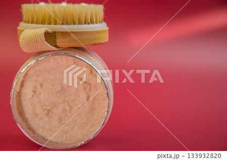 Exfoliating scrub jar with wooden brush on a bright red background, showcasing natural skincare products for beauty routines and self-care practices for wellness lovers 133932820