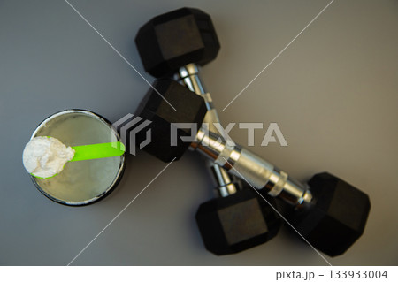 Fitness equipment includes black dumbbells next to a protein powder container with a green scoop, emphasizing health, exercise, and nutrition in a workout setting Fitness equipment includes black dumbbells next to a protein powder container with a green scoop, emphasizing health, exercise, and nutrition in a workout setting 133933004