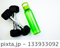 Dumbbells and a green water bottle are arranged on a white surface, symbolizing fitness and hydration, ideal for promoting a healthy lifestyle and exercise routines 133933092