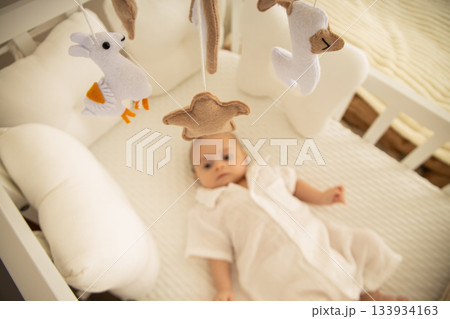 A serene baby resting in a cozy crib beneath soft toys A serene baby resting in a cozy crib beneath soft toys 133934163