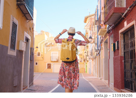 Woman in dress strolls through colorful streets of Spanish coastal town of La Vila Joiosa . sunny winter atmosphere highlights charm of Mediterranean architecture and quiet seaside life, back view Woman in dress strolls through colorful streets of Spanish coastal town of La Vila Joiosa . sunny winter atmosphere highlights charm of Mediterranean architecture and quiet seaside life, back view 133936286