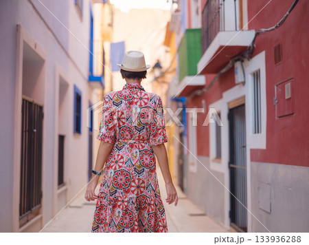 Woman in dress strolls through colorful streets of Spanish coastal town of La Vila Joiosa . sunny winter atmosphere highlights charm of Mediterranean architecture and quiet seaside life, back view Woman in dress strolls through colorful streets of Spanish coastal town of La Vila Joiosa . sunny winter atmosphere highlights charm of Mediterranean architecture and quiet seaside life, back view 133936288