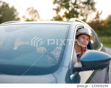 Traveler woman in red ethnic dress and hat drives car through scenic countryside of Croatia in summer. Surrounded by lush fields and forests, she enjoys peaceful and adventurous road trip 133936294