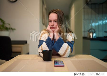 Female resting head on hands at kitchen table with mug and smartphone Female resting head on hands at kitchen table with mug and smartphone 133936462