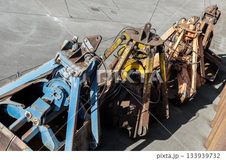 Row of heavy grab buckets rests on concrete surface, showing worn paint, rust textures and complex metal mechanics. Industrial tools create rugged atmosphere with strong sunlight and sharp shadows 133939732