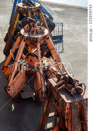 Row of heavy grab buckets rests on concrete surface, showing worn paint, rust textures and complex metal mechanics. Industrial tools create rugged atmosphere with strong sunlight and sharp shadows Row of heavy grab buckets rests on concrete surface, showing worn paint, rust textures and complex metal mechanics. Industrial tools create rugged atmosphere with strong sunlight and sharp shadows 133939743