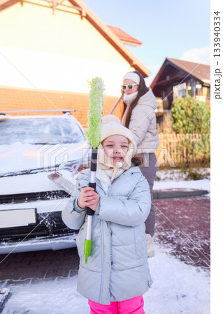 Child holds snow brush while woman clears car on a cold winter day in poland 133940334