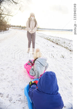 Mother Pulling Children on Sleds in Snowy Park on Winter Day near Water 133940349