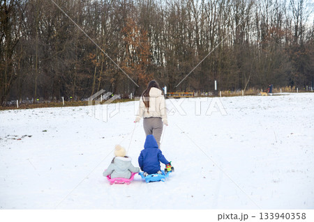 Woman pulls two children on sleds across a snowy field near a bare forest in Winter, enjoying an afternoon activity Woman pulls two children on sleds across a snowy field near a bare forest in Winter, enjoying an afternoon activity 133940358