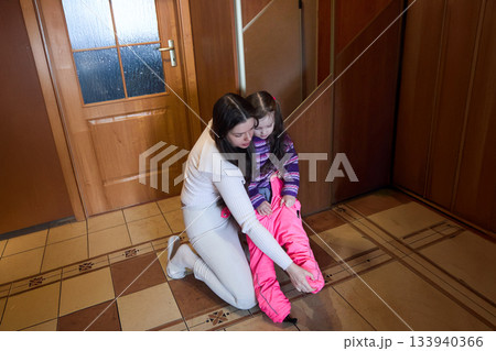Mother helps daughter put on snow pants indoors before going outside during the day near a door and hallway Mother helps daughter put on snow pants indoors before going outside during the day near a door and hallway 133940366