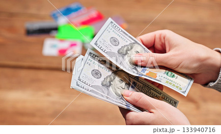 Woman counting US dollar bills, blurred credit cards in the background, symbolizing debt, loans, credit, financial illiteracy, and personal budgeting. Woman counting US dollar bills, blurred credit cards in the background, symbolizing debt, loans, credit, financial illiteracy, and personal budgeting. 133940716