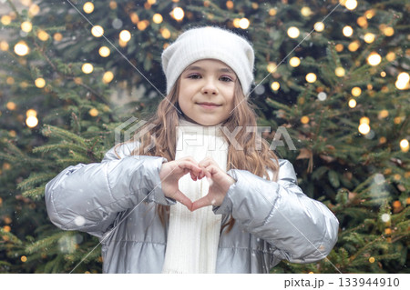 Girl in winter coat and scarf forms a heart with hands in a festive Christmas setting 133944910