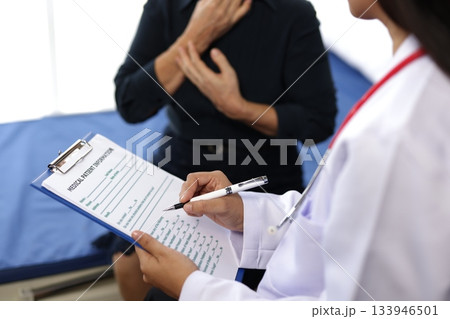 Asian female doctor wearing white coat and stethoscope consulting senior patient sitting on hospital bed, taking notes on clipboard in clinic room. Asian female doctor wearing white coat and stethoscope consulting senior patient sitting on hospital bed, taking notes on clipboard in clinic room. 133946501