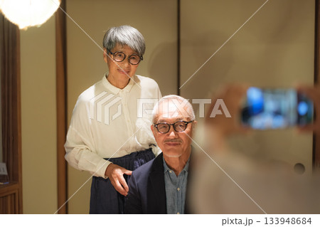 家族旅行の夕食で娘に還暦祝いの記念写真を撮ってもらうシニア夫婦 133948684