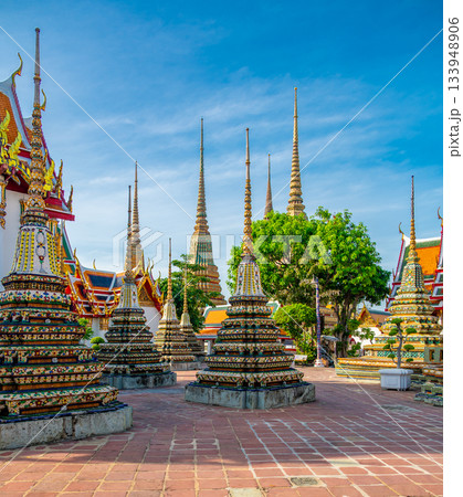 Exterior view of Wat Pho with richly decorated stupas covered in colorful floral ceramics and golden temple roofs shining under a clear blue sky on a bright sunny day in Bangkok. 133948906
