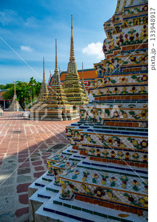 Exterior view of Wat Pho with richly decorated stupas covered in colorful floral ceramics and golden temple roofs shining under a clear blue sky on a bright sunny day in Bangkok. 133948917