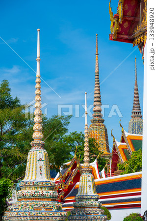 Exterior view of Wat Pho with richly decorated stupas covered in colorful floral ceramics and golden temple roofs shining under a clear blue sky on a bright sunny day in Bangkok. 133948920