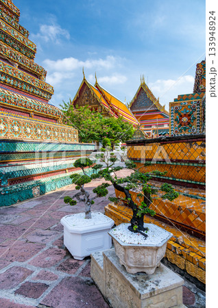 Exterior view of Wat Pho with richly decorated stupas covered in colorful floral ceramics and golden temple roofs shining under a clear blue sky on a bright sunny day in Bangkok. 133948924