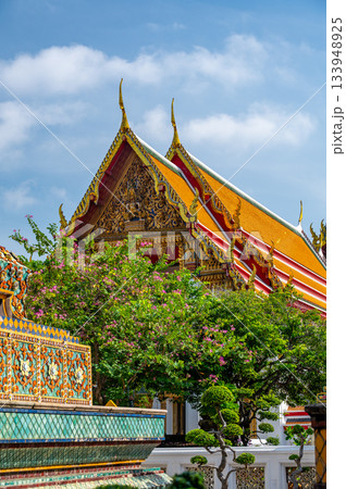 Exterior view of Wat Pho with richly decorated stupas covered in colorful floral ceramics and golden temple roofs shining under a clear blue sky on a bright sunny day in Bangkok. 133948925