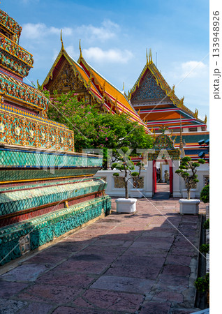 Exterior view of Wat Pho with richly decorated stupas covered in colorful floral ceramics and golden temple roofs shining under a clear blue sky on a bright sunny day in Bangkok. 133948926