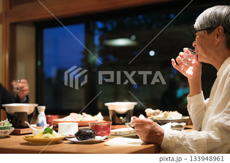 シニア夫婦が還暦祝いの旅行で旅館の会席料理をゆったり味わう夕食シーン 133948961