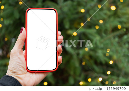 Smartphone with empty blank screen in womans hand against Christmas tree with light garland. Screen display for Festive Xmas Holiday Background. Space for text. Christmas advertising, smartphone app Smartphone with empty blank screen in womans hand against Christmas tree with light garland. Screen display for Festive Xmas Holiday Background. Space for text. Christmas advertising, smartphone app 133949700
