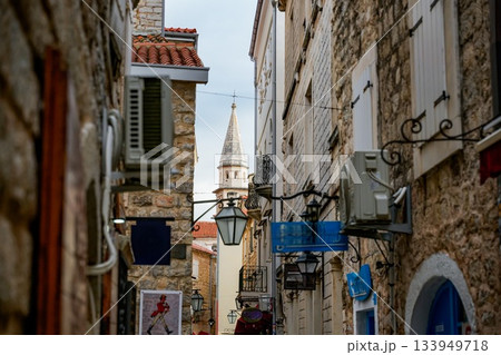 Commercial photo of a narrow stone street with wall-mounted lanterns leading to the bell tower of the Cathedral of Saint John the Baptist (Catholic) in Budva, Montenegro. Medieval architecture 133949718