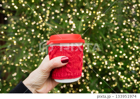 Hand holding red coffee cup with knitted sleeve in front of bright Christmas tree lights. Cozy festive winter mood with warm bokeh. Cozy outdoor coffee. Hand holding red coffee cup with knitted sleeve in front of bright Christmas tree lights. Cozy festive winter mood with warm bokeh. Cozy outdoor coffee. 133951974