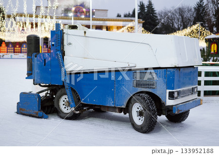 Ice resurfacer machine in blue and white smoothing the surface of an outdoor skating rink decorated with bright holiday lights. Ice resurfacer machine in blue and white smoothing the surface of an outdoor skating rink decorated with bright holiday lights. 133952188
