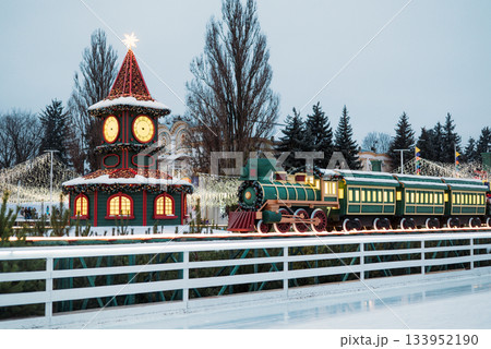 Festive Christmas village display with a toy clock tower and a green holiday train next to a decorated outdoor ice skating rink. 133952190