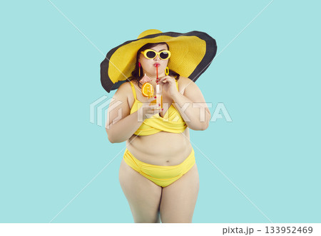 Carefree young plus size woman wearing beachwear drinking cocktail on blue studio background. 133952469