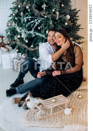 Couple enjoying cozy moment by Christmas tree, sharing laughter and love while surrounded by holiday decorations and a playful pet Couple enjoying cozy moment by Christmas tree, sharing laughter and love while surrounded by holiday decorations and a playful pet 133954057