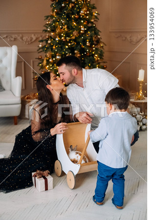 Family enjoys a cozy Christmas moment with a wooden toy cart, a Christmas tree, and a child observing a playful kitten 133954069