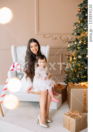 Mother and daughter enjoy a festive moment by the Christmas tree with presents in a cozy living room decorated for the holidays 133954110