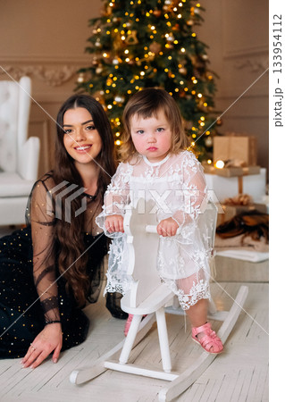Young girl in a beautiful dress plays on a rocking horse with her mother nearby, festive decorations adding warmth to the room during the holiday season 133954112