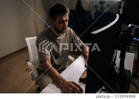 Man concentrating on desktop computer, working from home office on freelance project, demonstrating dedication and productivity in informal remote office environment. Man concentrating on desktop computer, working from home office on freelance project, demonstrating dedication and productivity in informal remote office environment. 133954909