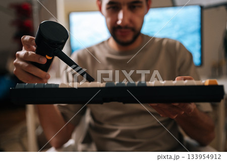 Selective focus view of bearded male cleaning computer mechanical keyboard at home using electric air duster, performing essential maintenance for technology devices and hardware hygiene. 133954912