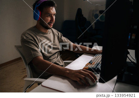Focused young man wearing headphones and casual t-shirt, concentrating playing video game on desktop computer, enjoying immersive entertainment during leisure time at home in dark room. Focused young man wearing headphones and casual t-shirt, concentrating playing video game on desktop computer, enjoying immersive entertainment during leisure time at home in dark room. 133954921