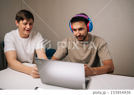 Two male friends enjoying competitive video gaming on laptop sitting at desk at home, connecting and having fun listening with headphones and concentrating using game controller. Two male friends enjoying competitive video gaming on laptop sitting at desk at home, connecting and having fun listening with headphones and concentrating using game controller. 133954936