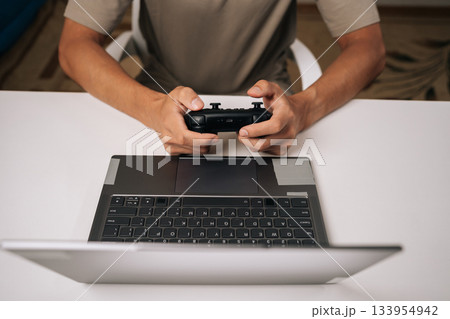 Top view of gamer male gaming on laptop computer using black wireless gamepad controller, enjoying modern online entertainment during leisure time, hands holding joystick device over keyboard. Top view of gamer male gaming on laptop computer using black wireless gamepad controller, enjoying modern online entertainment during leisure time, hands holding joystick device over keyboard. 133954942