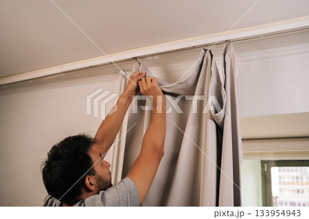 Man reaching up to attach light-colored curtain to white track rod on ceiling, focusing on setting up home decor in apartment, representing domestic life. Concept of household cleanliness. Man reaching up to attach light-colored curtain to white track rod on ceiling, focusing on setting up home decor in apartment, representing domestic life. Concept of household cleanliness. 133954943