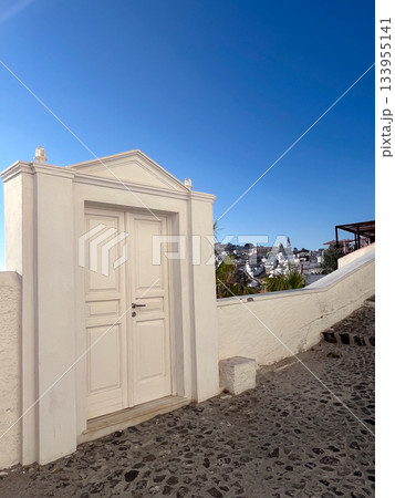 Greek island scene with a classic white door opening onto a cobblestone street, offering a picturesque village view and the Aegean sea beyond under a clear blue sky. 133955141