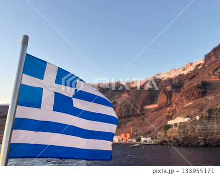 Greek flag waving proudly on a boat sailing near the island cliffside houses of Santorini, Aegean sea, basking in the glow of a warm sunset light 133955171