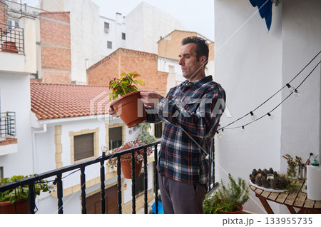 A man wearing a plaid shirt adjusts a potted plant on a balcony, adding greenery to an urban outdoor space. String lights create a warm, cozy mood. 133955735