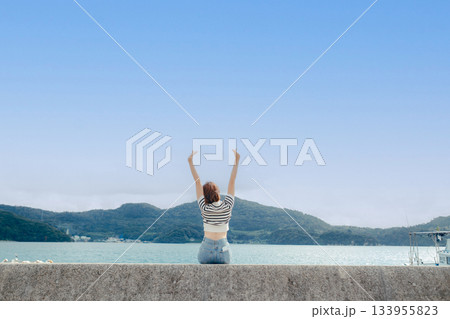 海を眺めてひと休みする20代女性。旅先で感じる風と光のエモーショナルな時間 133955823