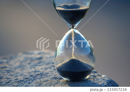 Elegant hourglass with black sand trickling down, set against a serene blue background 133957239