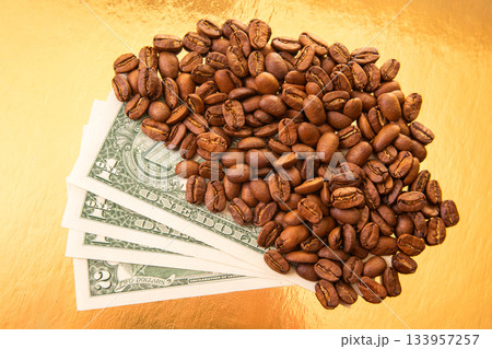 Coffee beans spill over U.S. dollar bills on a reflective golden surface Coffee beans spill over U.S. dollar bills on a reflective golden surface 133957257