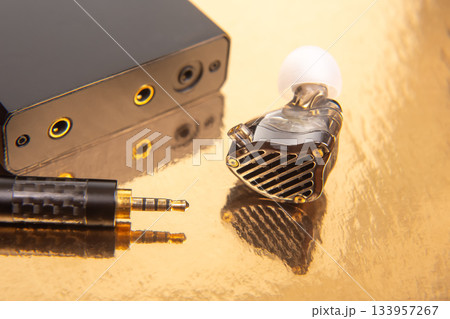 Close-up of an earbud, a gold-plated 3.5mm plug, and a music player on a glossy golden background 133957267