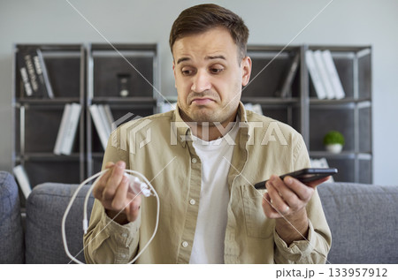 Upset frustrated man holding broken mobile phone and charger with cable in hands 133957912