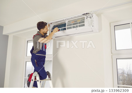 Professional electrician man with screwdriver maintaining modern air conditioner indoors. Professional electrician man with screwdriver maintaining modern air conditioner indoors. 133962309